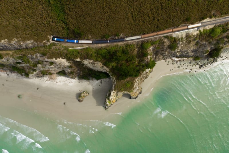 Aerial view of The Seaside train traveling along an Otago coastal cliff above a sandy beach and turquoise ocean.