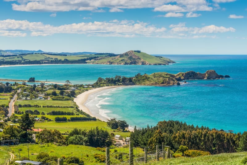 An elevated Otago coastal view shows a curving white sand beach meeting turquoise ocean water, with green hills and a blue sky above.