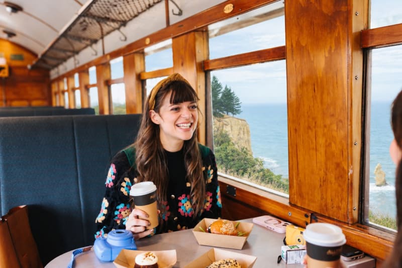 A smiling woman in The Seasider train carriage holds a coffee cup, with coastal views visible through the window.