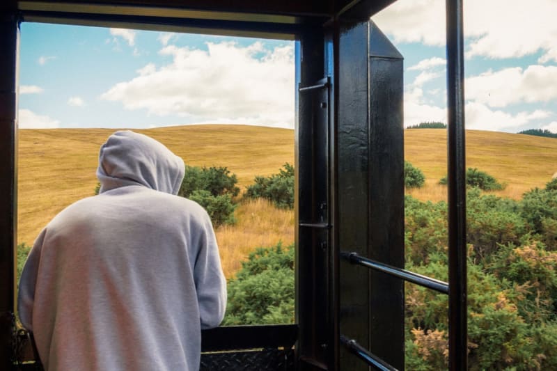Two smiling women lean out of the Taieri Gorge train window, looking at a smartphone while traveling through a mountainous  Otago landscape.