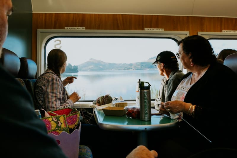 A smiling woman wearing a floral-patterned sweater holds a coffee cup at a table inside The Seasider wooden train car with ocean views.