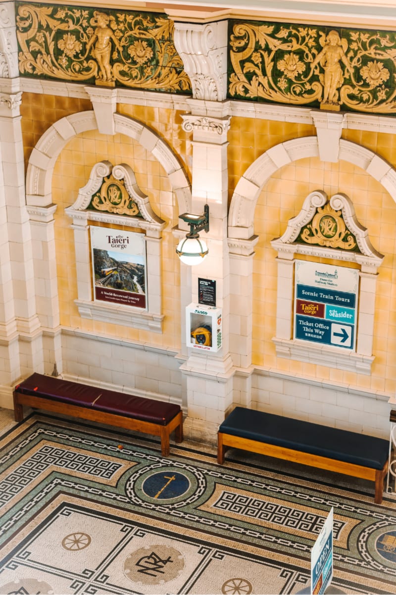 High-angle shot of an ornate yellow and white Dunedin Railway Station interior with tiled walls, arched panels, wooden benches, and a mosaic floor.
