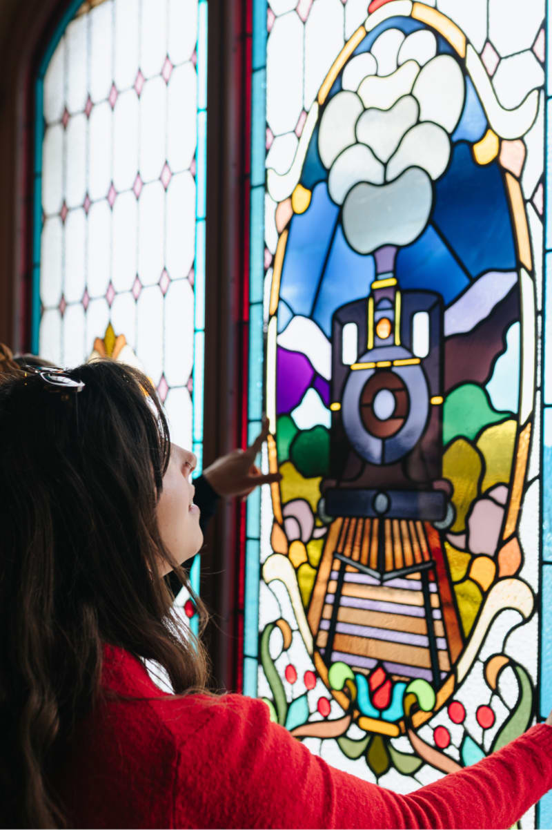 A woman in a red sweater points at a colorful stained-glass window depicting a steam train on tracks.