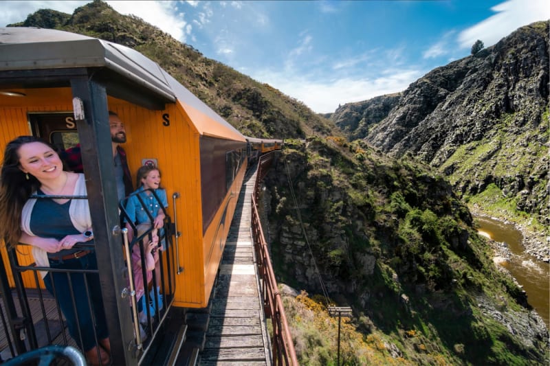 Passengers look out from the open-air viewing platform of the Taieri Gorge train traveling along a steep mountain gorge.