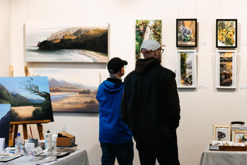 Two visitors view framed landscape and floral paintings hanging on a white gallery wall at the Dunedin Railway Station.