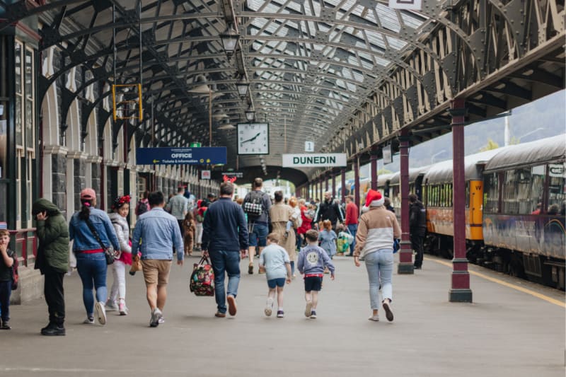 People walking along a busy platform at the Dunedin railway station, next to a parked train.