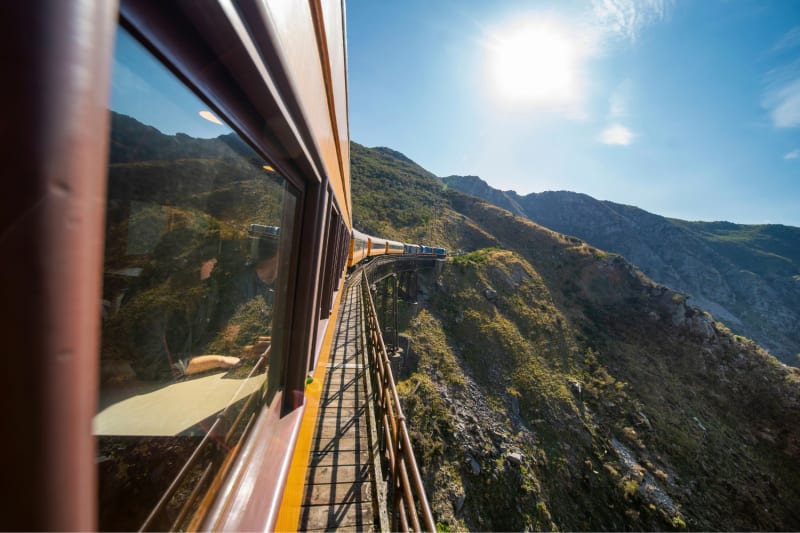 The Taieri Gorge train travels along a winding mountain railway track under a bright sun.