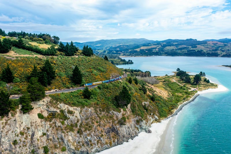 The Seasider train travels along a narrow causeway beside a body of water within Dunedin with a rolling green hill in the background.