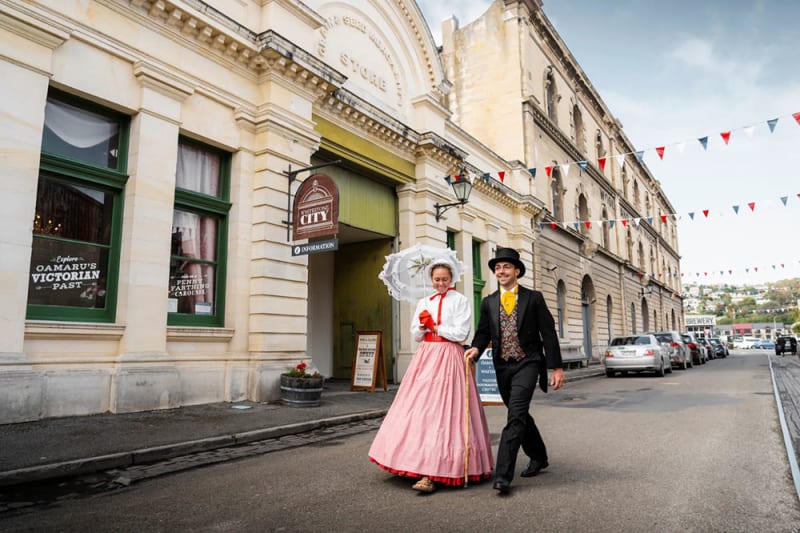 A man and woman in Victorian-era clothing walk down a historic cobblestone Oamaru, NZ street lined with old stone buildings.