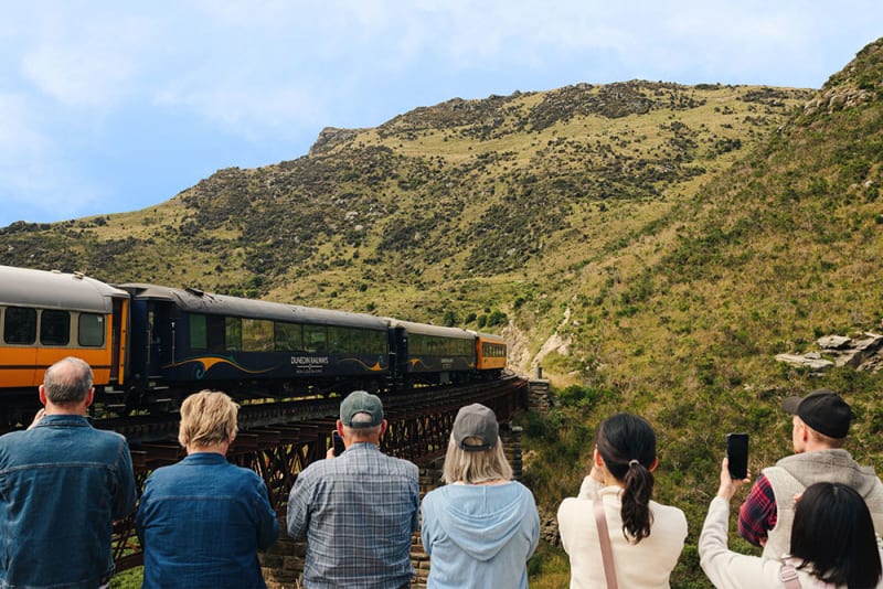An aerial view of The Taieri Gorge train traveling along a curving track through th lush, green Taieri valley beside th winding Taieri river.