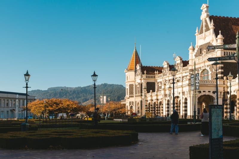 An aerial view of the historic Dunedin Railway Station in New Zealand, featuring formal gardens and a seagull flying in the foreground.