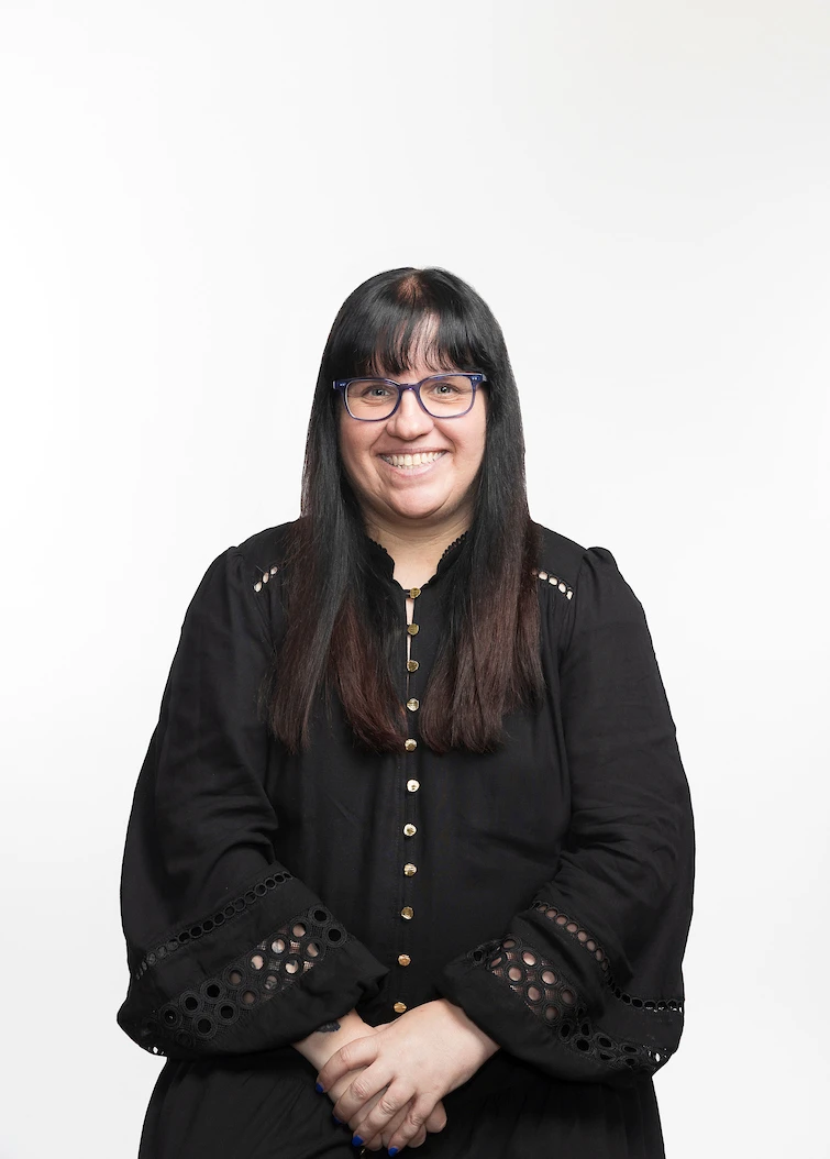 Portrait of Christine McNamara smiling with long dark hair and blue glasses, wearing a black blouse against a plain white background.