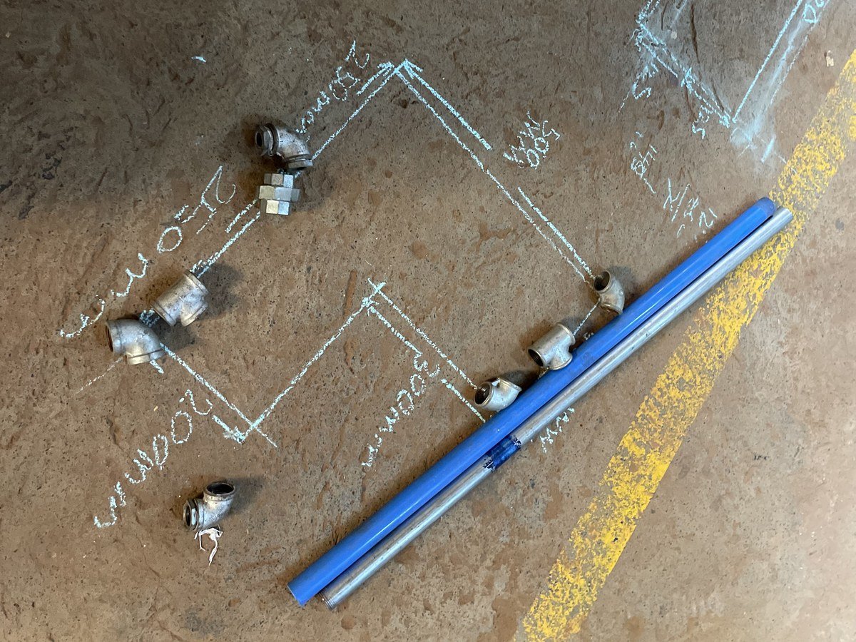 Chalk pipe layout drawn on a workshop floor with galvanised elbow fittings and two lengths of pipe laid beside it