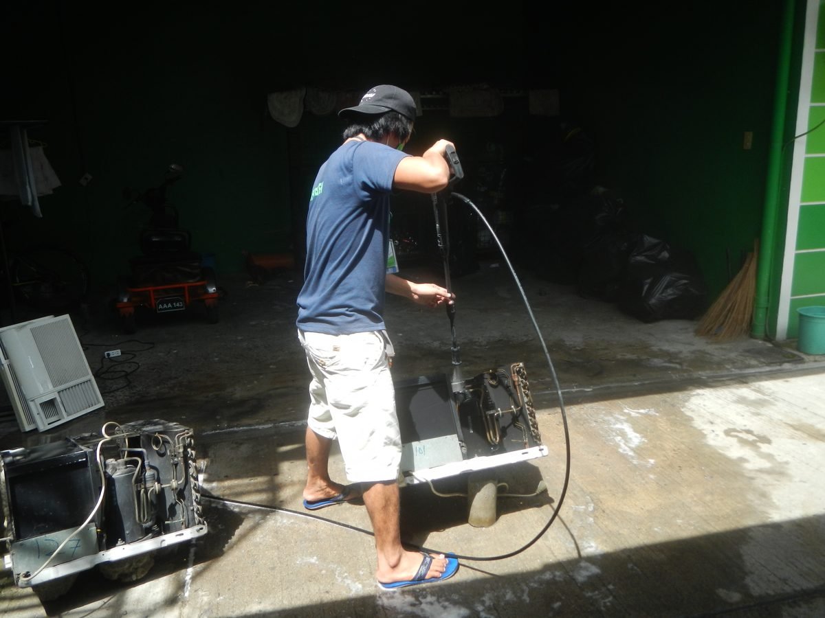 A technician using a pressure washer on an air conditioner unit laid on a concrete floor in a yard