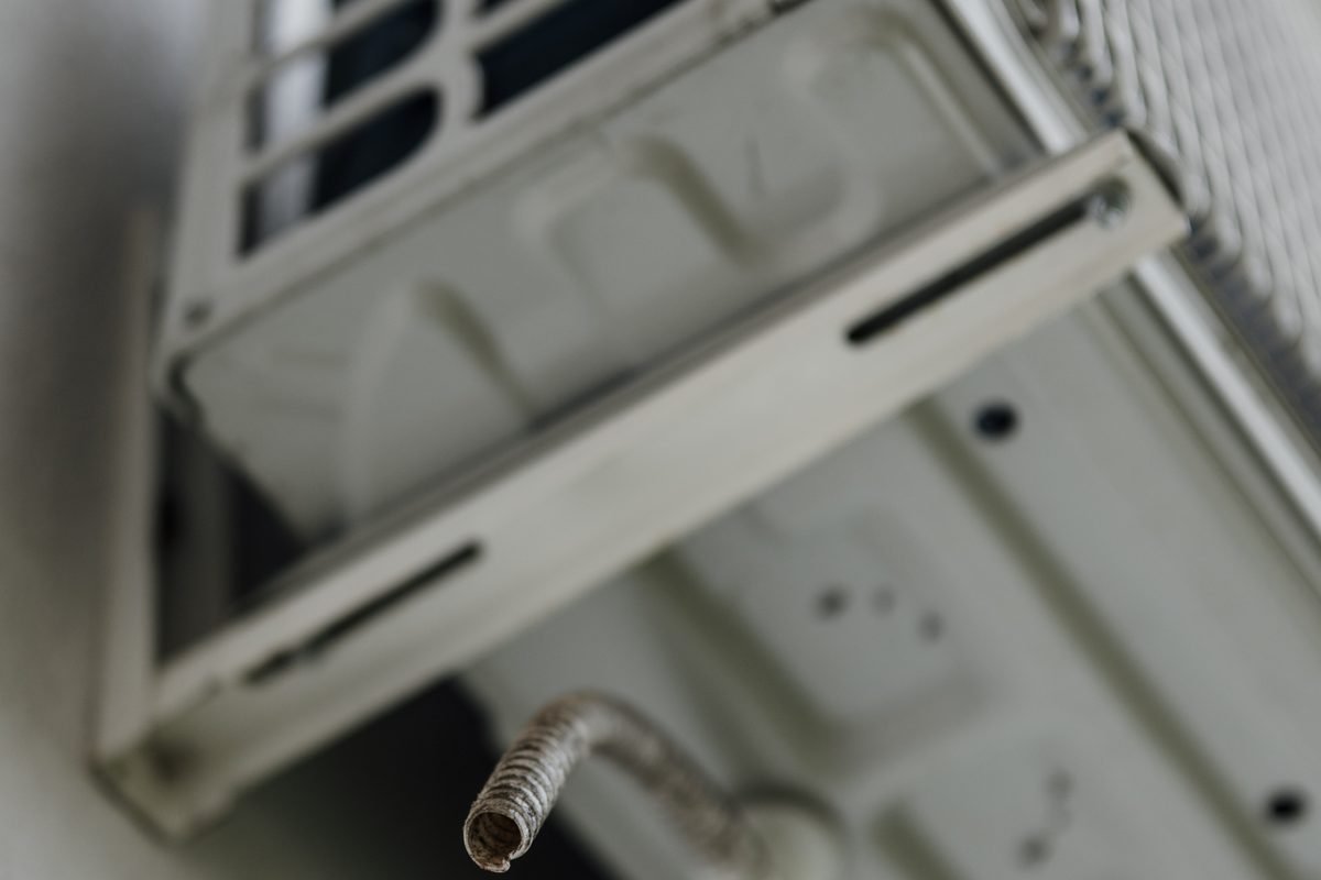 Underside of an outdoor air conditioner unit on wall brackets, with a short drain hose hanging below