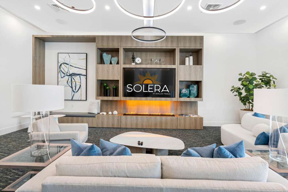 Modern lounge area with white sofas, decorative accents, and a wall-mounted TV with an electric fireplace at Solera at Avalon Trails in Delray Beach, Florida