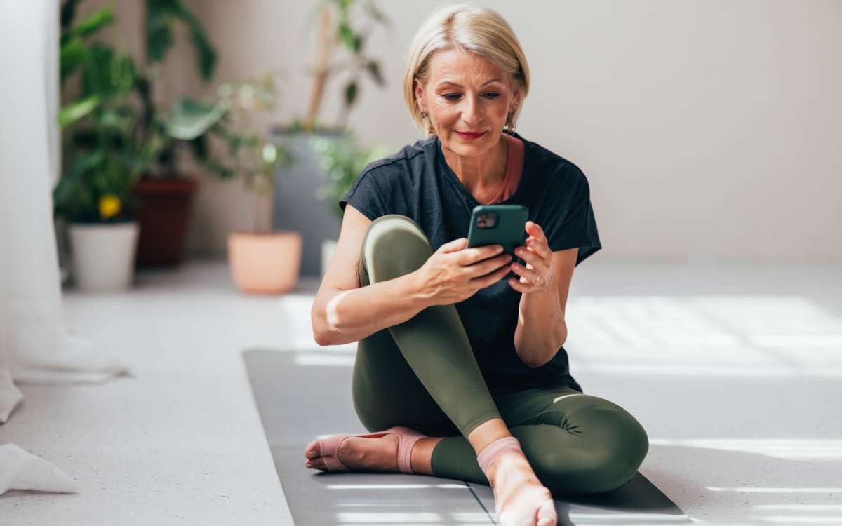 Woman sitting on yoga mat, using smartphone, surrounded by potted plants in bright room at Alamitos on 4th in Long Beach, California 