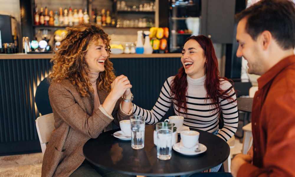 Three friends laughing and enjoying coffee at a modern café table with drinks and pastries near Everett at Port Wentworth in Port Wentworth, Georgia
