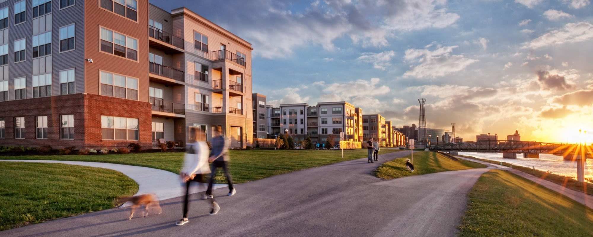 Two people walking a dog along a path near modern apartment buildings at sunset at Water Street Apartments in Dayton, Ohio