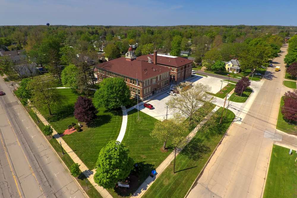Historic building surrounded by green lawns and trees, with a paved pathway and parking area at Cavalier Greene in Corunna, Michigan
