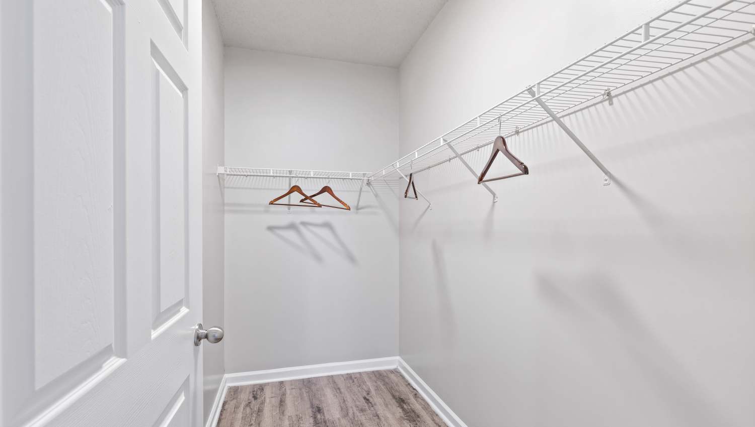 Empty walk-in closet with white walls, wooden floor, wire shelves on three sides, and several wooden hangers. Partially visible door on the left