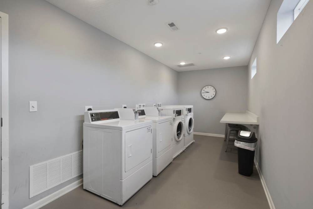 Bright laundry room with multiple machines, folding table, and trash bin against light gray walls at Wheatland Crossing II in Columbus, Ohio