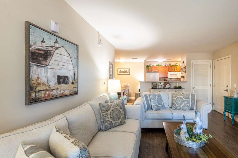 Cozy living room with wood-style flooring, light-colored sofas, and a fully-equipped kitchen in background at White Oak Crossing in Knoxville, Tennessee
