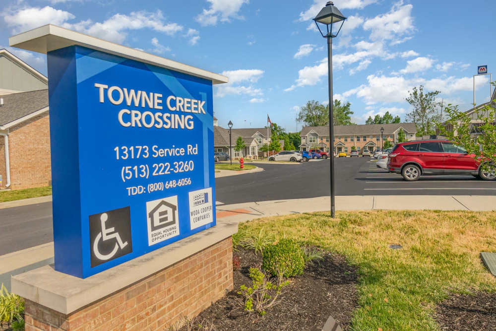 Community sign with accessible features and landscaped entrance, featuring parking lot and nearby residences at Towne Creek Crossing in Walton, Kentucky