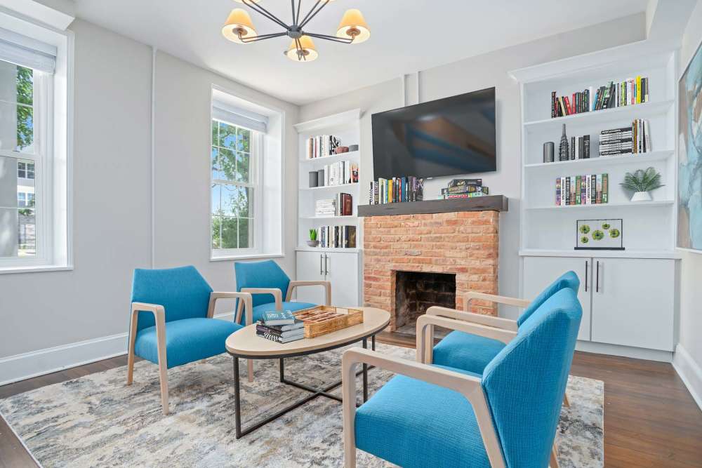 Bright lounge with blue chairs, coffee table, bookshelves, and a brick fireplace. Large windows provide natural light at Crestwood Manor in Frederick, Maryland