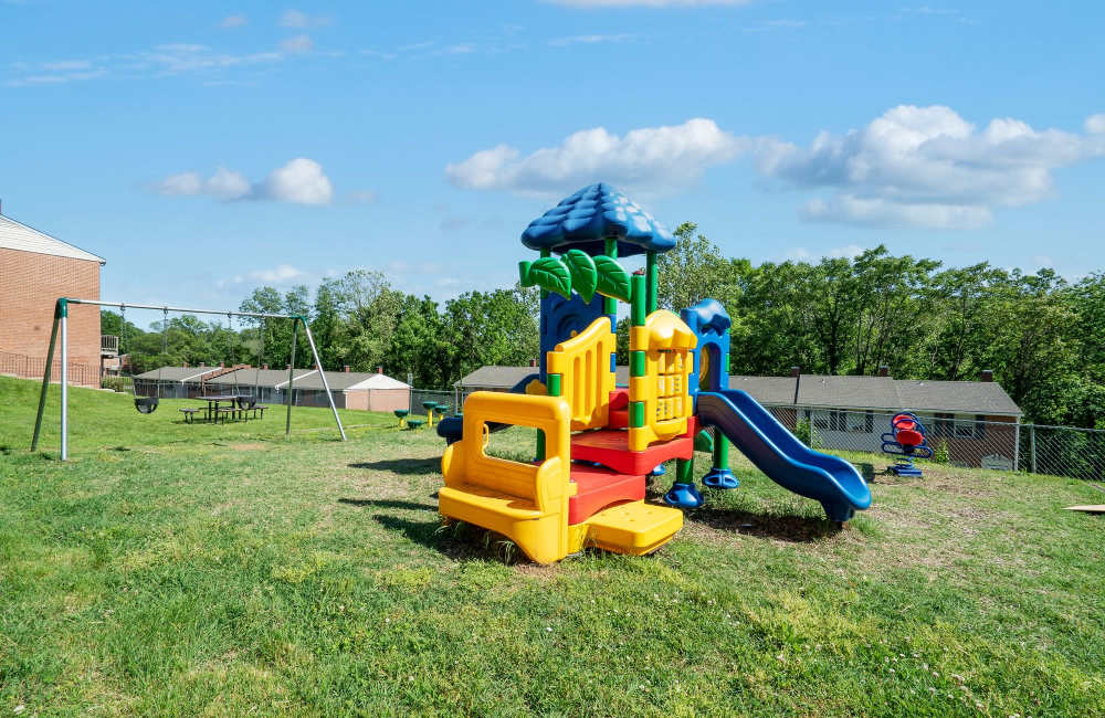 Colorful playground with slides and swings in grassy area, surrounded by trees and apartment buildings at Fairway Ridge in Baltimore, Maryland