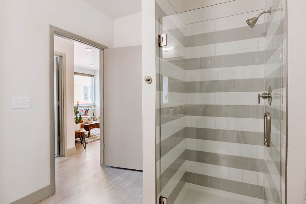 Modern glass shower with striped tile design and sleek fixtures, bright open layout visible beyond at Viña Apartments in Denver, Colorado
