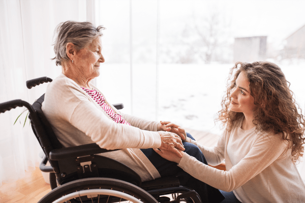 Granddaughter lovingly holding wheelchair-bound grandmother's hands while looking up affectionately