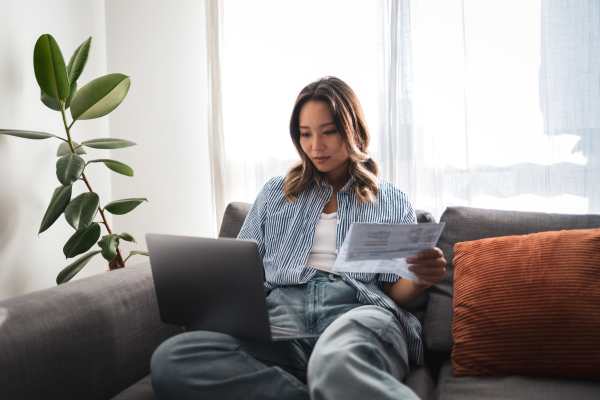 Woman sitting on couch using laptop while holding documents with indoor plants nearby at Valley Brook Townhomes in Rochester, New York