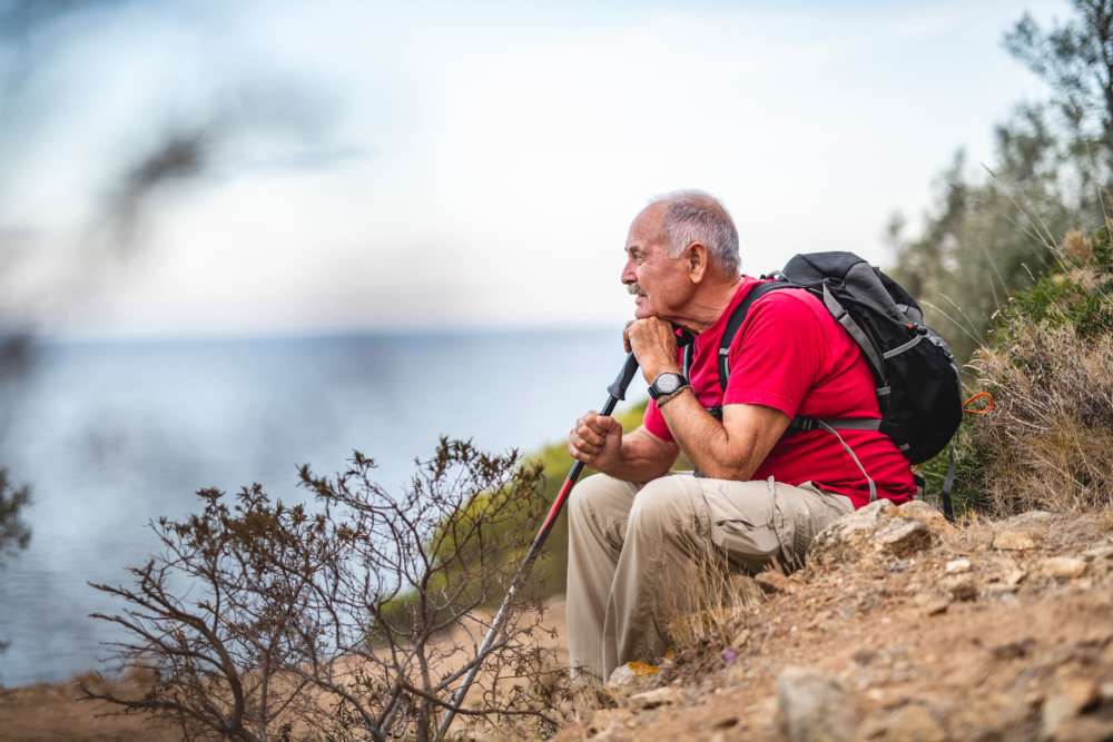 Senior man resting on a rocky trail overlooking the water, holding a hiking pole and wearing a backpack at Alamitos on 4th in Long Beach, California