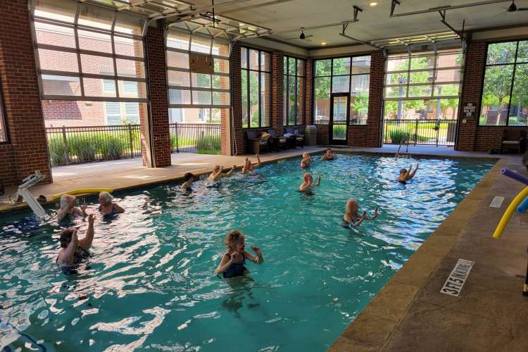 Seniors participating in a water aerobics class in a community pool, with lush greenery and trees visible outside through large windows at Watermere at Frisco in Frisco, Texas.