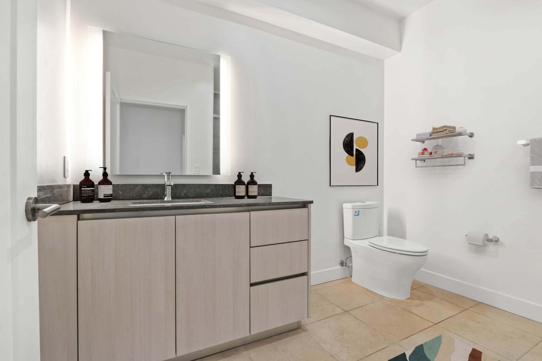 Modern bathroom with light wood vanity, large mirror, white toilet, wall shelf, and abstract artwork at Luxe Villas in Brentwood, California 