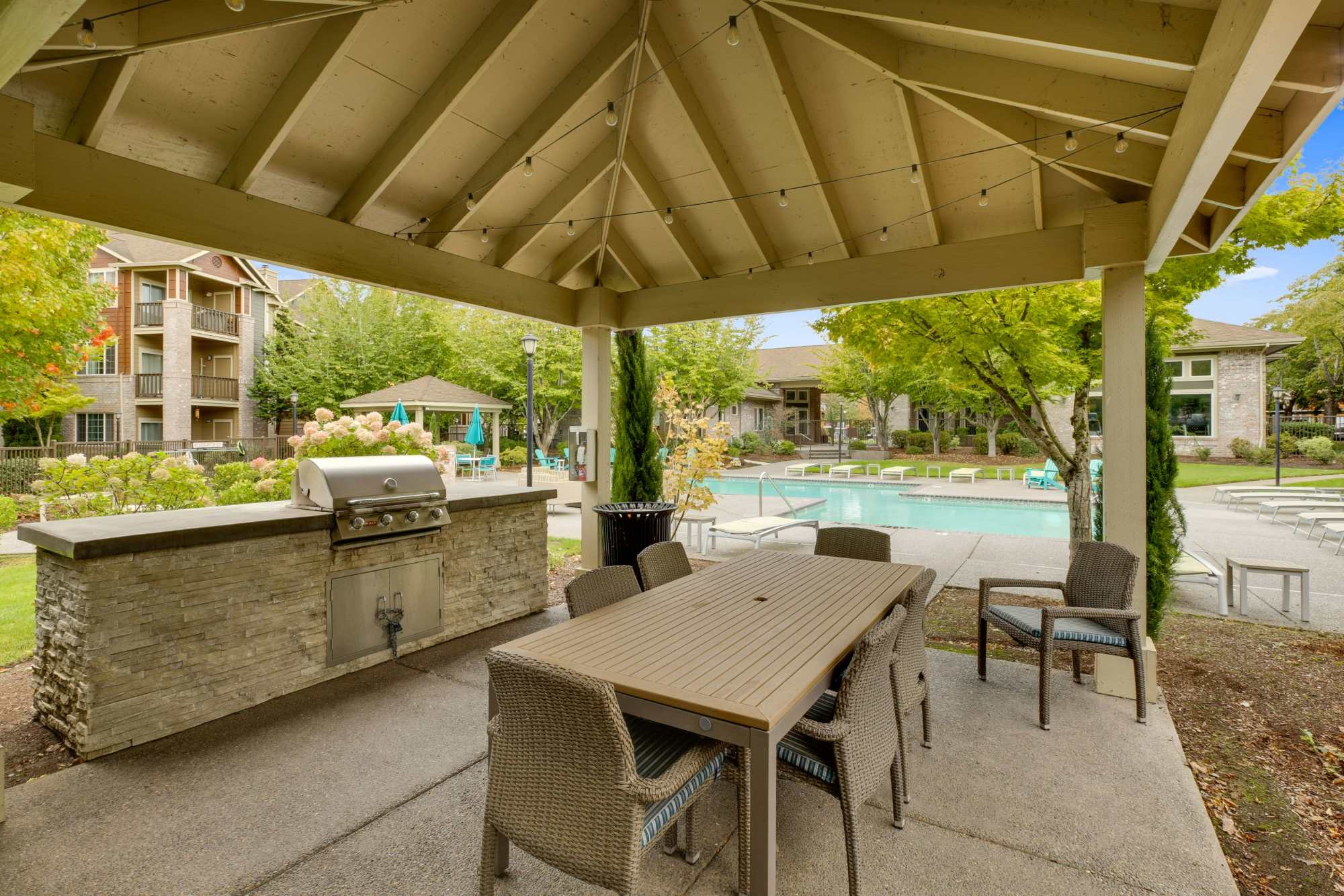 A covered patio area by the pool with a large wooden table and rope lights on the ceiling at The Grove at Orenco Station in Hillsboro, Oregon