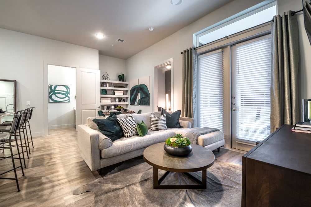 North Dallas, TX, Apartments Near Addison Airport - Cornell Midtown - Stylish Living Room With Sectional Sofa, Round Coffee Table, Open Shelving, and Glass Patio Doors With Blinds And Drapes.