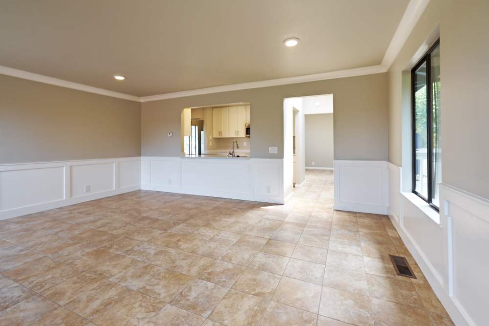 Empty room with beige walls, tiled flooring, white wainscoting, crown molding, large window, recessed lighting, and a pass-through to the kitchen. at Santa Cruz Coast Homes in Santa Cruz, California