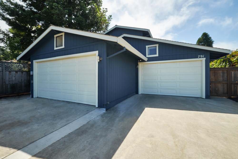 Blue house with two white garage doors, concrete driveway, surrounded by trees and a wooden fence, with a logo in the top right corner. at Santa Cruz Coast Homes in Santa Cruz, California