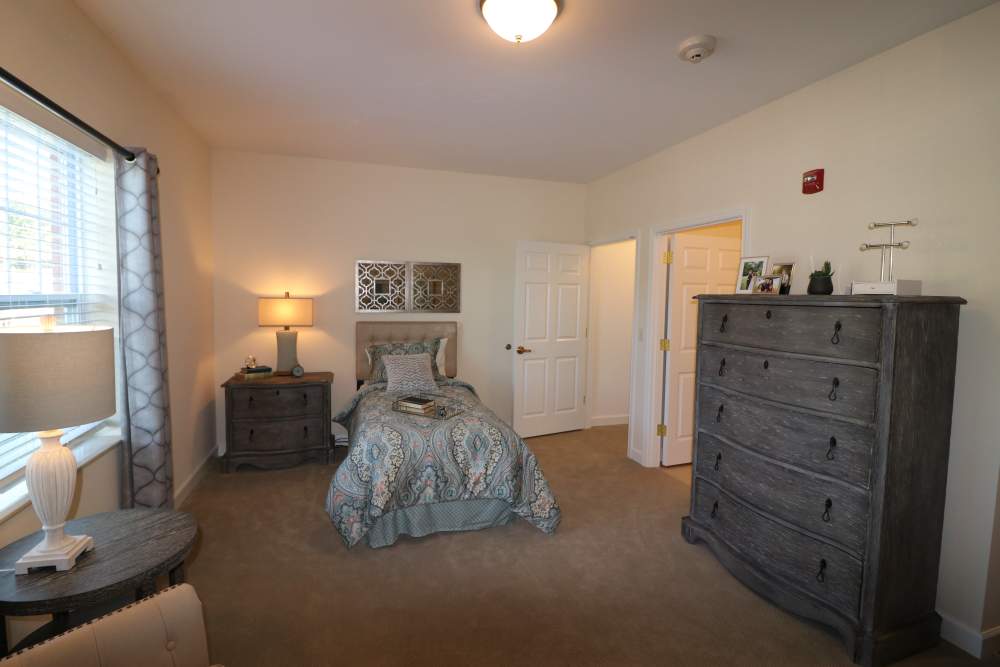 Bedroom with window, night stand with a lamp and storage space at The Harmony Collection at Roanoke - Assisted Living in Roanoke, Virginia
