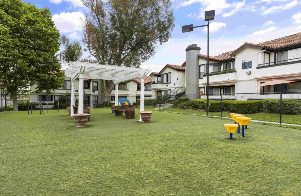 One Bedroom Apartments in Corona CA - Brookwood Villas - Dog Park with Manicured Grass, Fence, Agility Equipment, and Benches Under a Pergola