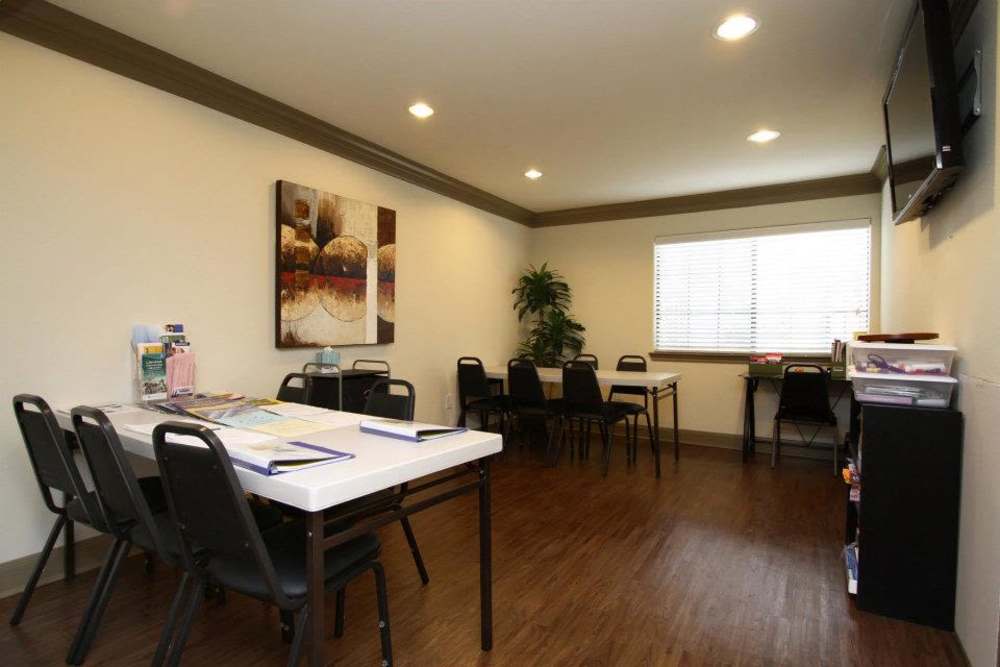 Modern community room with wood-style flooring, tables, chairs, and a large window with blinds at Villa Del Prado in Houston, Texas