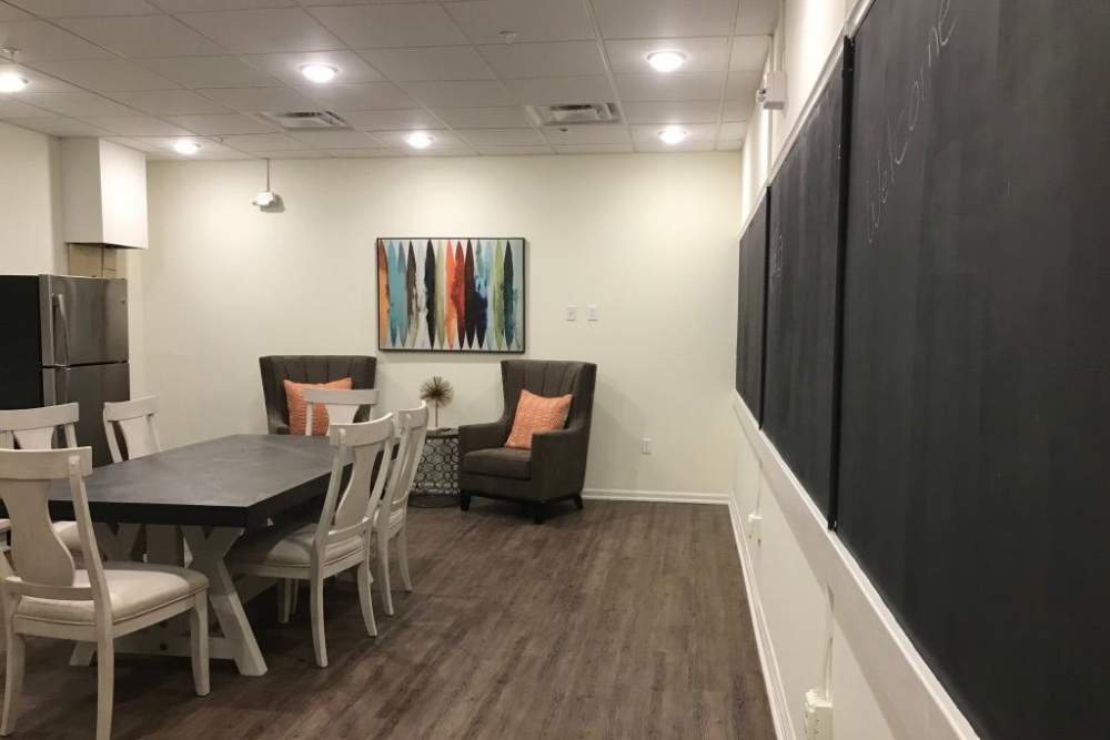 Modern community room with wood-style flooring, large dining table, comfortable seating, and chalkboard walls at Cavalier Greene in Corunna, Michigan