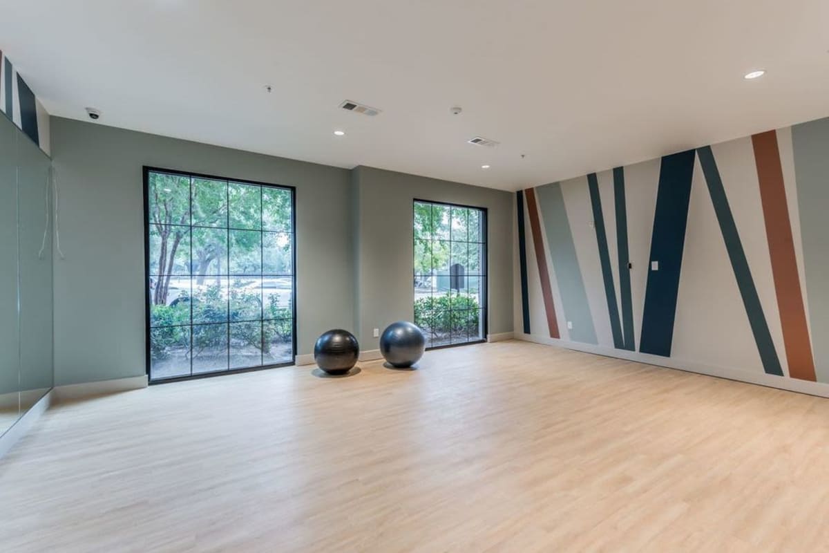 A spacious exercise or yoga studio with a light wooden floor and two large windows allow natural light to enter, offering a view of greenery outside at Villas At Bunker Hill, Houston, Texas