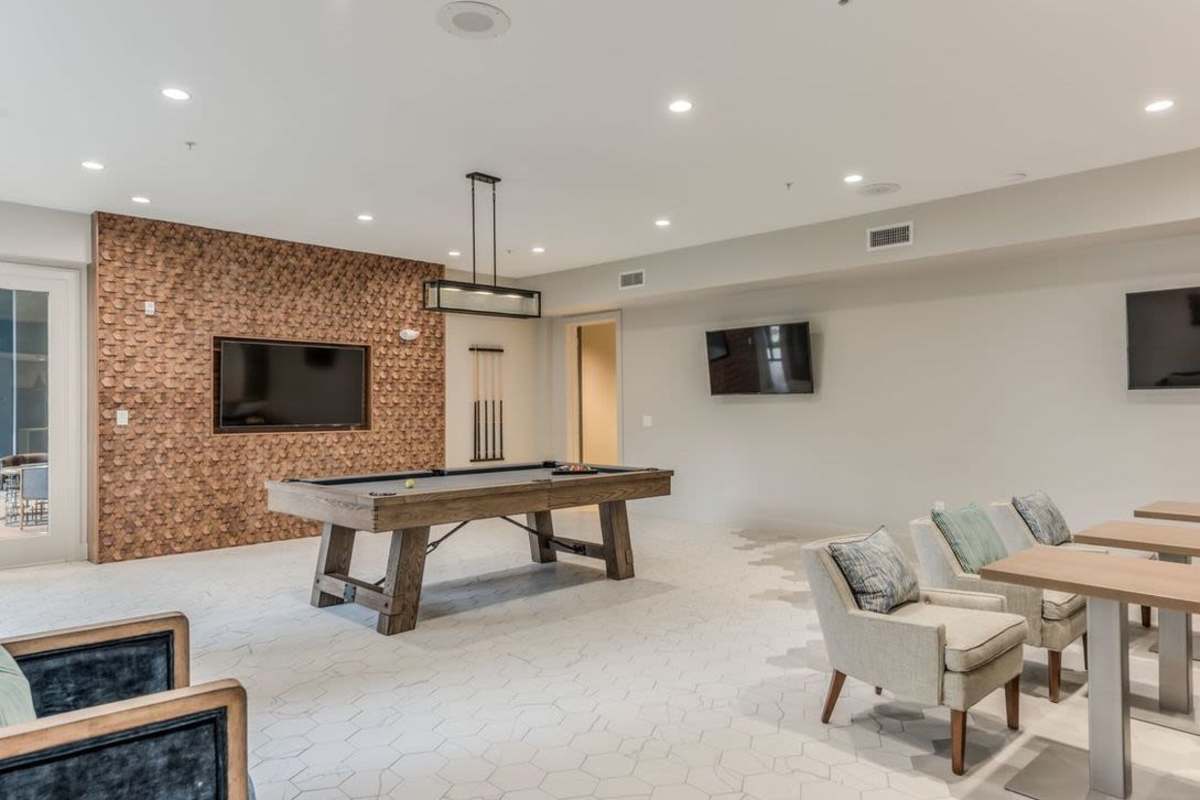 A stylish communal lounge area featuring a billiard table in the center with one wall is adorned with a decorative texture, and there are two flat-screen TVs mounted on the walls at Villas At Bunker Hill, Houston, Texas