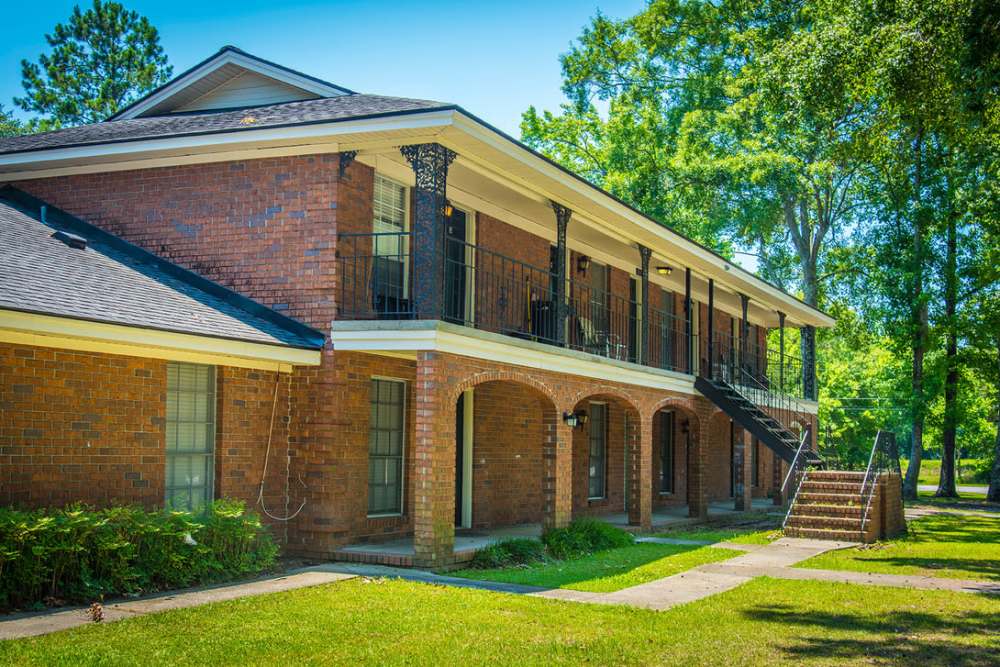 Two-story brick apartment building with arched porches and a staircase leading to the second floor, surrounded by lush green trees and grass at Magnolia Downs in Charleston, South Carolina
