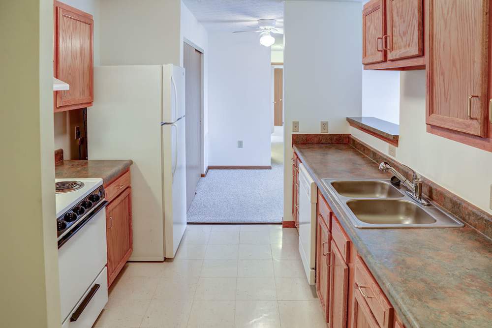 Galley kitchen with white appliances, wooden cabinets, and double sink leading to carpeted living area at Heritage Greene in Washington Court House, Ohio