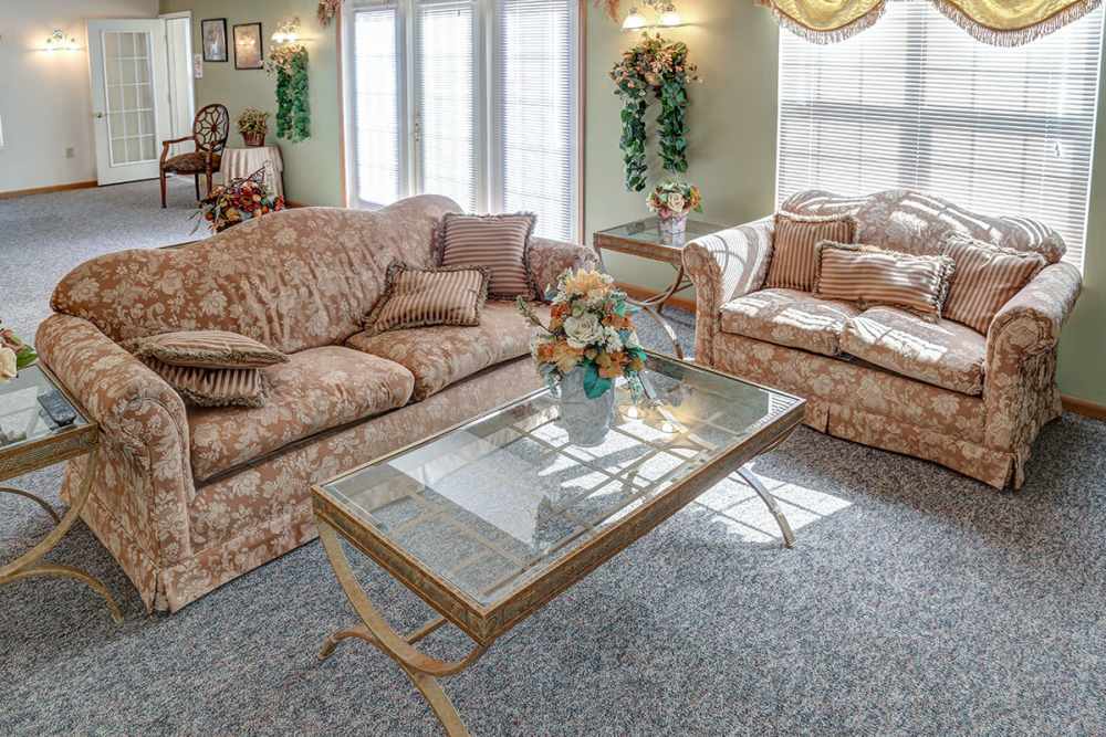 Cozy living room with floral couches, glass coffee table, and natural light from large windows at Heritage Greene in Washington Court House, Ohio