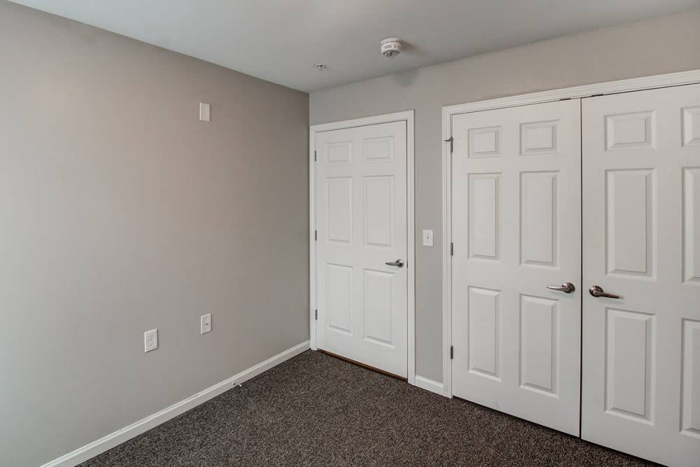 Empty room with beige walls and two white doors, carpeted flooring, and a smoke detector on the ceiling at Northside Drive Apartments in Crossville, Tennessee